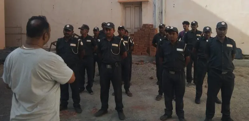A man in a grey shirt briefing a group of guards in uniform at an outdoor site